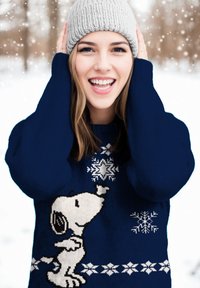Maglione blu navy con un grande design di Snoopy lavorato a maglia e fiocchi di neve bianchi. Il modello indossa un berretto lavorato a maglia grigio chiaro, sorridente.