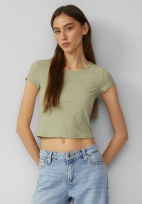 Olivinvihreä, poimutettu, lyhythihainen crop-top, jossa on pyöreä kaula-aukko, yhdistettynä vaaleansinisiin korkeavyötäröisiin farkkuihin. Sujuva rakenne, tiivis malli.