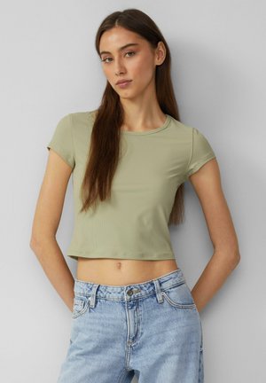 Olivovo zelený, žebrovaný crop top s krátkym rukávom a okrúhlym výstrihom, skombinovaný so svetlomodrými džínami s vysokým pásom. Hladká textúra, priliehavý strih.