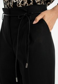 Zbliżenie na osobę w czarnych spodniach z cienkim paskiem związanym w kokardę oraz ręką spoczywającą w kieszeni.