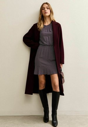 Cappotto lungo bordeaux sopra un vestito a pois bianco e nero, stivali neri al ginocchio, con in mano una piccola borsa strutturata, sfondo neutro.