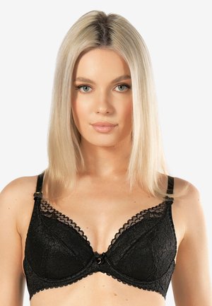 Blondynka o prostych włosach, ubrana w czarny koronkowy biustonosz, zwrócona przodem na jasnym, jednolitym tle.