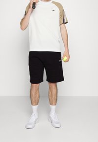 Vit t-shirt med kort ärm och beige detaljer samt mönstrade ärmar, tillsammans med svarta shorts. Modellen håller en tennisracket och en boll.
