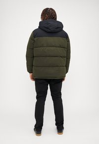 Veste matelassée en vert olive avec une capuche noire et une partie supérieure noire. Présente un design matelassé horizontal et une coupe ample. Associée à un pantalon sombre.