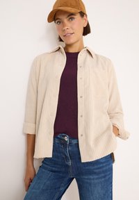 Camicia in corduroy beige con chiusura a bottoni e maniche arrotolate, indossata sopra un maglione viola a coste, abbinata a jeans blu. Cappellino marrone indossato.