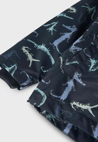Tessuto di giacca blu navy con motivo a coccodrillo in verde e azzurro chiaro su tutta la superficie. Presenta polsini a coste e una superficie liscia.
