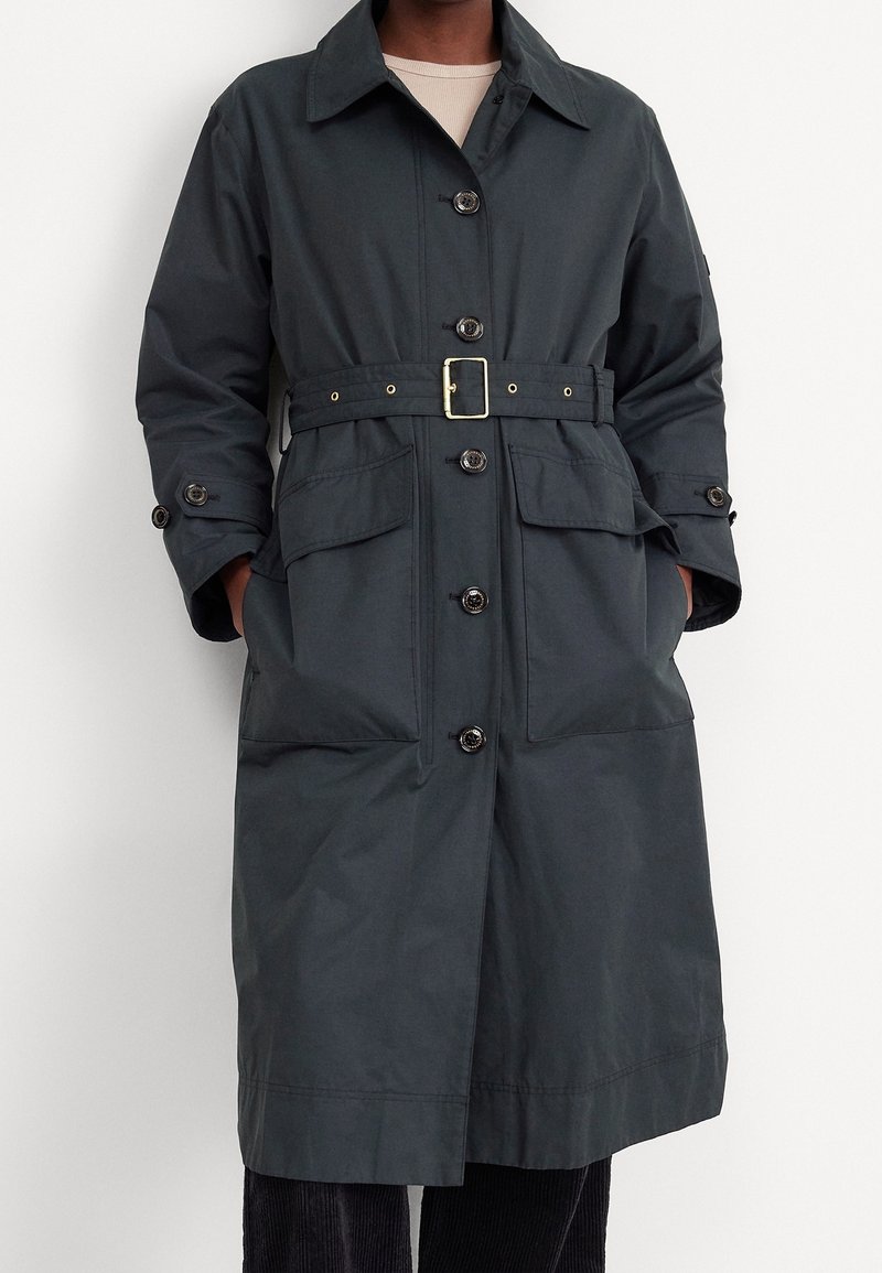 Trench-coat gris foncé ceinturé avec de gros boutons et poches à rabat à l'avant, porté sur une chemise claire et un pantalon foncé.