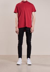 Camiseta de algodón roja con cuello alto y texto "HUNDRED", combinada con pantalones negros ajustados y zapatillas blancas, sobre un fondo neutro.