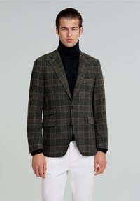 Blazer de cuadros gris y verde con dos bolsillos frontales, cierre de un botón y tejido texturizado, combinado con un cuello de tortuga negro y pantalones blancos.