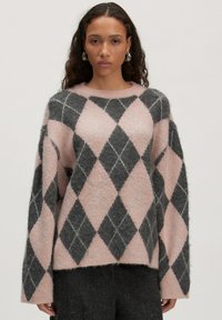Vrouw in een fuzzy roze en donkergrijze kraag met lange mouwen en zwarte broek, staand tegen een effen achtergrond.