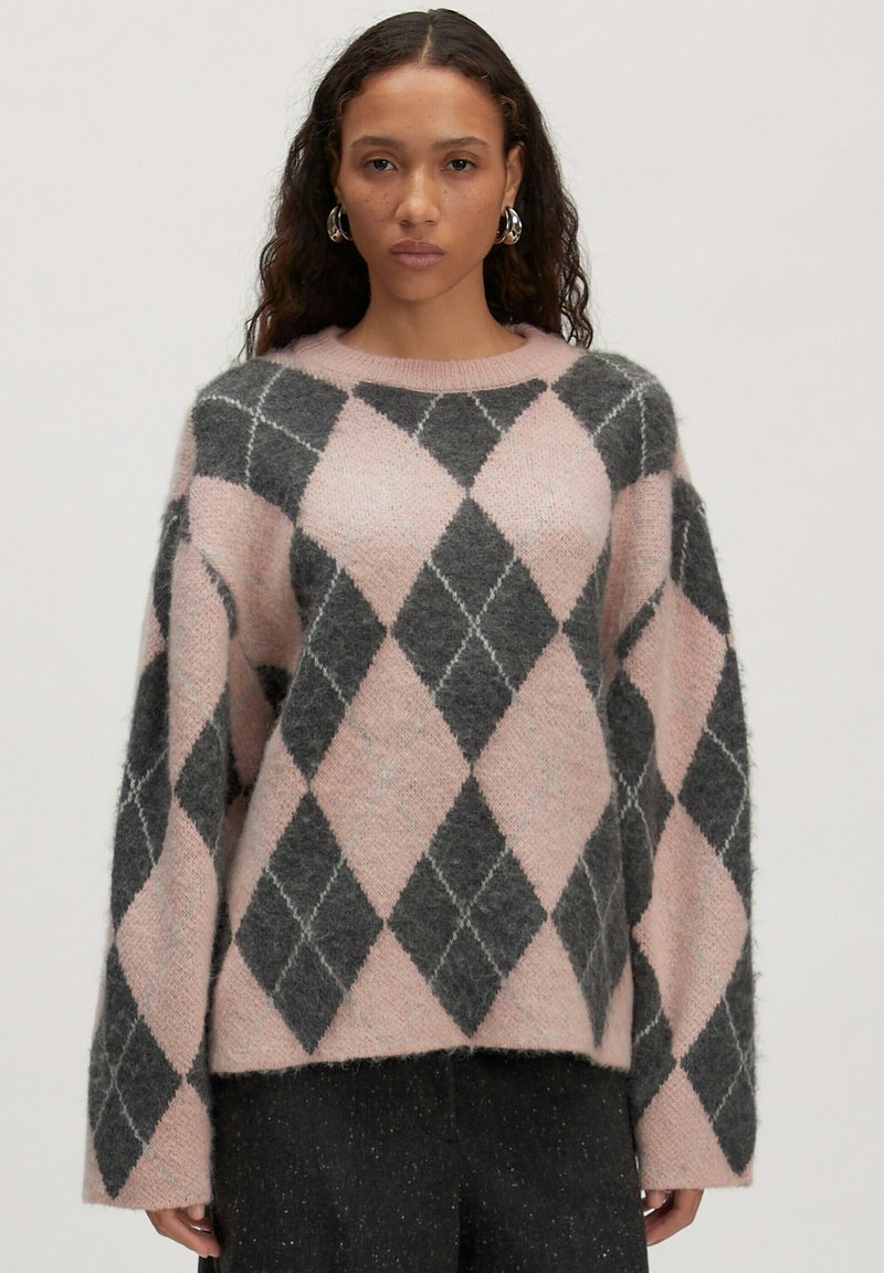 Vrouw in een fuzzy roze en donkergrijze kraag met lange mouwen en zwarte broek, staand tegen een effen achtergrond.