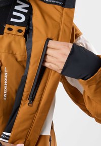 Giacca marrone resistente all'acqua con accenti neri e bianchi, dotata di una tasca con zip e fodera interna a contrasto. Tessuto morbido.