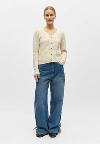 Femme debout, portant un cardigan en maille crème à boutons, un jean bleu ample avec des liens aux ourlets, et des chaussures noires, sur un fond blanc uni.