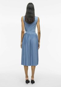Modrá plisovaná midi šaty s priliehavým telom a širokým pásom, s hladkou textúrou a rozšírenou sukňou. Nosia sa s čiernymi topánkami na blokovom opätku.