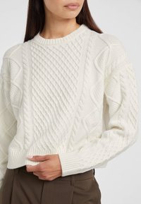 Maglione color crema a treccia con un motivo testurizzato, colletto e polsini a coste, e una vestibilità morbida, abbinato a pantaloni marroni.