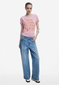 Camiseta de algodón rosa con impresión en naranja que dice "See You by The Sea", combinada con vaqueros de mezclilla azul claro de gran tamaño con costuras y bolsillos distintivos.