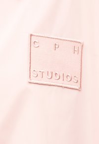 Ορθογώνιο ροζ υφασμάτινο πατς με κεντημένο το κείμενο "CPH STUDIOS" ραμμένο πάνω σε φόντο ανοιχτού ροζ υφάσματος.