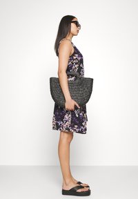 Kvinde iført en sort blomsterkjole og sorte sandaler, der holder en stor flettet sort håndtaske, stående i profil mod en hvid baggrund.