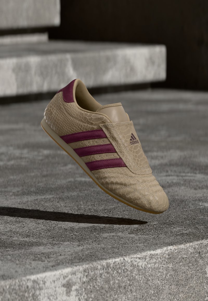 Baskets couleur beige avec un motif texturé imitation peau de serpent, ornées de rayures et d'accents violets. Semelle en caoutchouc et logo Adidas sur le côté.