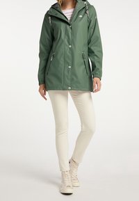 Personne portant un imperméable à capuche vert, un t-shirt rayé blanc, un pantalon skinny crème et des baskets blanches montantes, debout sur un fond uni.