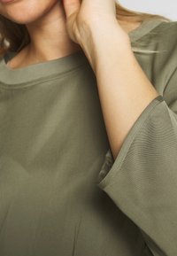 La blusa verde oliva presenta un collo rotondo e un tessuto morbido e fluente. Le maniche sono a tre quarti con orlo liscio.