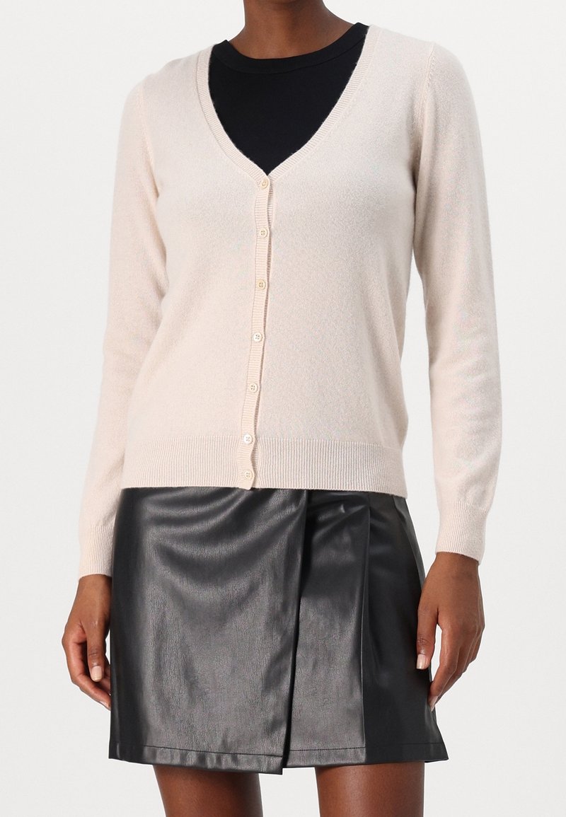 Femme portant un cardigan beige clair boutonné sur un haut noir, associé à une jupe en cuir noire avec un pli central à l'avant.
