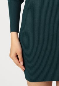 Rochie tricotată ribbed în nuanță teal, cu mâneci lungi, având o siluetă ajustată și un material texturat, evidențiind linii verticale subtile și o potrivire strânsă.