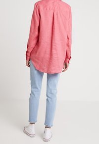 Chemise en lin rose à manches longues avec un ourlet courbé, portée sur un jean bleu clair. Comprend des poignets à boutons et un pli au dos pour faciliter les mouvements.