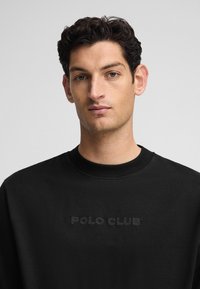 Sudadera negra con cuello redondo y un logo en relieve de "POLO CLUB" en el pecho. La tela parece suave con un corte clásico.