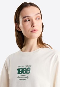Mujer joven con cabello castaño mirando hacia la derecha, vestida con una camiseta blanca con texto verde que dice "The North Face 1966 Est. Berkeley California".