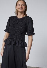 Sort top med sammensamlet tekstur og korte puf ærmer, med en flæset kant. Matchet med en let sort nederdel, sat mod en neutral baggrund.