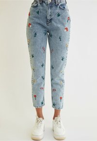 Jasnoniebieskie dżinsy z denimu z kwiatową haftem w kolorach czerwonym, żółtym i białym, charakteryzujące się wysokim stanem i zwężanymi nogawkami. Noszone z białymi trampkami.