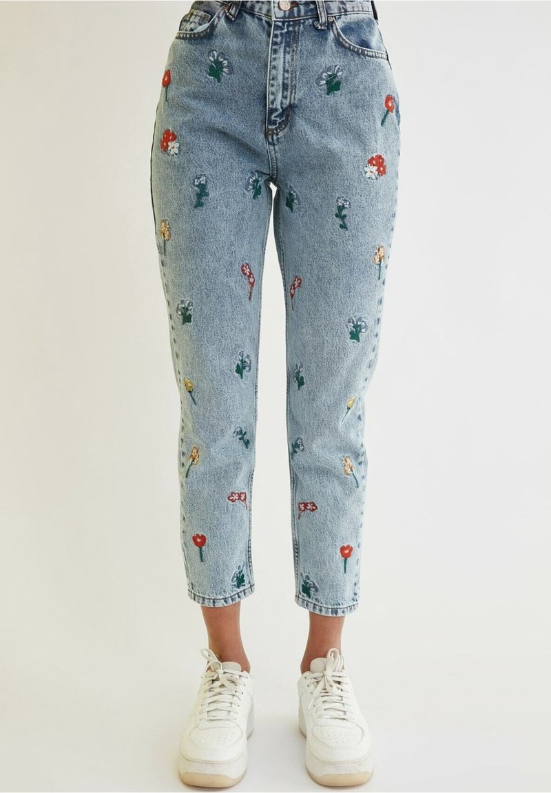 Jasnoniebieskie dżinsy z denimu z kwiatową haftem w kolorach czerwonym, żółtym i białym, charakteryzujące się wysokim stanem i zwężanymi nogawkami. Noszone z białymi trampkami.