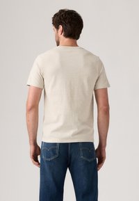 Mann, der mit dem Rücken zur Kamera steht, trägt ein schlichtes, beigefarbenes T-Shirt mit kurzen Ärmeln und blaue Jeans vor einem neutralen Hintergrund.