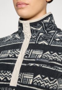 Svart og hvit mønstrete fleece-pullover med en beige krage, med knapper og Columbia-logo på nedre venstre side.