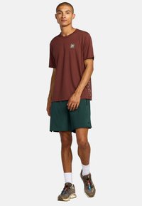 Bruine t-shirt met korte mouwen en een klein logo op de borst, gecombineerd met donker groene shorts, witte sokken en bruine sportschoenen.