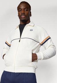 Hvit zip-up jakke med marineblå og oransje striper, med stående krage, ribbede mansjetter og en logo på brystet. Glatt stofftekstur.