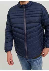 Uomo che indossa una giacca trapuntata blu navy con zip frontale e jeans blu, in piedi con le braccia rilassate su uno sfondo semplice.