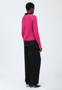 Rosa gestrickter Pullover mit strukturierten Ärmeln und geripptem Saum, kombiniert mit lockeren schwarzen Hosen und High Heels, von hinten präsentiert.
