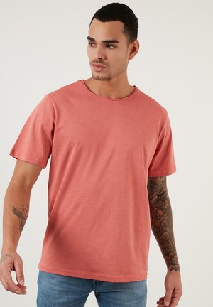 Tricou scurt, de culoare corai, cu mânecă scurtă și guler rotund, realizat dintr-un material moale, având o croială lejera și o textură minimă.