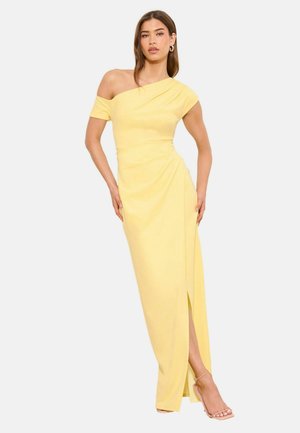 Kobieta w żółtej sukni sięgającej ziemi, z odsłoniętym jednym ramieniem, rozcięciem i sandałami na wysokim obcasie, stojąca na jednolitym tle.