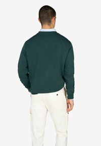 Felpa verde scuro con collo rotondo e polsini a costine, abbinata a pantaloni cargo beige chiaro con tasche laterali e dettaglio con bottone.