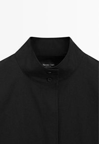 Schwarzes Hemd mit Stehkragen, ausgestattet mit einem Knopfverschluss und sauberer Nahtführung. Hergestellt aus einem glatten, leichten Stoff. Markenlabel sichtbar.