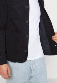 Svart kordjacka med fyra knappar och bröstficka, med en mjuk textur och blått foder. Bärs över en vit skjorta.