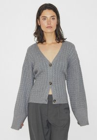 Grå stickad kofta med V-hals, ribbad textur och överdimensionerade ärmar. Har fem bruna knappar längs fram.