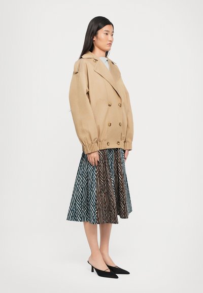Beige dubbele-breasted jas met uitlopende mouwen, gecombineerd met een A-lijn rok met een patroon van blauwe, bruine en witte chevron strepen. Zwarte pumps met hak.