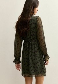 Mujer con cabello largo castaño, vistiendo un vestido verde oliva transparente con estampado floral negro, con cintura ajustada y puños con volantes, de espaldas.