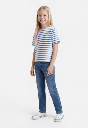 Ung jente står, iført en hvit og blåstripet kortermet skjorte med sløyfedetalj, blå jeans og hvite joggesko, smilende.