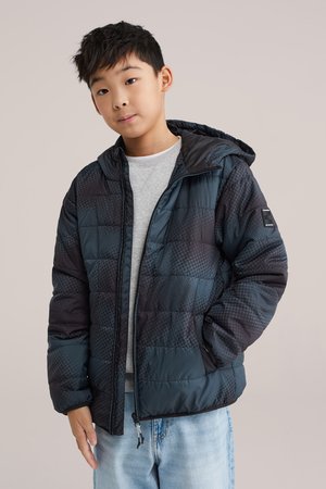 Băiat purtând o geacă matlasată cu model în albastru închis și negru, cu glugă, pulover gri deschis și blugi albastru deschis, stând pe un fundal neutru.