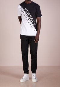 Czarny i biały t-shirt z diagonalnym wzorem, na którym widnieje tekst "VERSACE COLLECTION" napisany dużą czcionką. Połączony z czarnymi spodniami i białymi sneakersami.